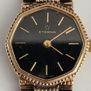 May include: A gold-tone wristwatch with a black face and gold hands. The watch face is octagonal and has the word "ETERNA" printed in gold letters. The watch band is a gold-tone metal link band.