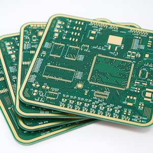 May include: A stack of four green circuit boards with intricate gold and white electronic components. The boards have rounded corners and a gold border, creating a layered effect. The boards are arranged on a white background.