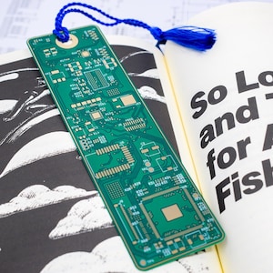 May include: A green circuit board bookmark with a blue tassel. The bookmark is lying on a book with the text "So Long, and Thanks for All the Fish".