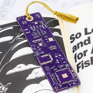 May include: A purple circuit board bookmark with a gold tassel, placed in an open book. The bookmark features a detailed electronic component design. The book's page displays the text "So Long, and Thanks for All the Fish."