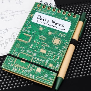 Pode incluir: Um pequeno caderno verde com uma faixa elástica preta e um rótulo branco que diz "Daily Notes". O caderno está sobre uma superfície preta com uma caneta verde.