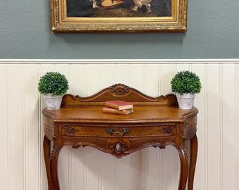 May include: A wooden console table with curved legs, a single drawer, and ornate carvings. Two small potted green plants sit on the ends of the surface, flanking two stacked books. Above the table hangs a framed painting of three chickens.