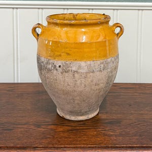 May include: A vintage, two-toned ceramic jar with a rounded body and two small handles. The upper portion is a warm yellow, while the lower half is a textured, neutral gray. This antique vessel is likely used for storage.