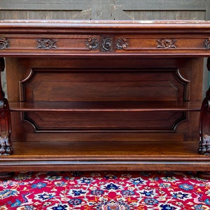 May include: Antique dark wood console table with ornate carvings and lion's paw feet. The table has two shelves and a marble top.