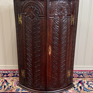 May include: A dark brown, antique corner cabinet with intricate carved leaf and floral designs. The cabinet has two doors with brass hinges and a small keyhole. The cabinet is curved and stands on a patterned rug.