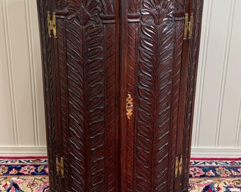 May include: A dark brown, antique corner cabinet with intricate carved leaf and floral designs. The cabinet has two doors with brass hinges and a small keyhole. The cabinet is curved and stands on a patterned rug.