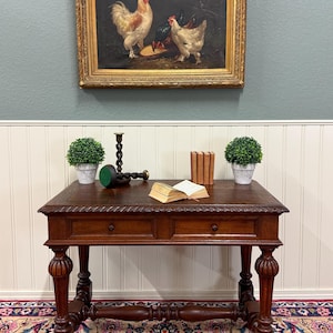 May include: A dark brown wooden desk with two drawers, turned legs, and a carved edge. On top sits an open book, three small books, two green potted plants, and a black twisted metal candlestick. A framed painting of chickens hangs above.
