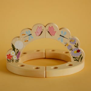 May include: A wooden, circular toy with a scalloped top, decorated with painted flowers, butterflies, dragonflies, and bees. The toy is made of light-colored wood and is composed of four interlocking pieces. The background is a solid yellow.