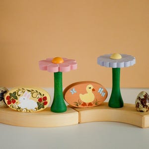 May include: Easter decorations made of wood. Two flower-shaped ornaments with pink and light blue petals, green stems, and yellow centres. Two painted wooden egg-shaped ornaments with a rabbit and duck, and two speckled eggs.
