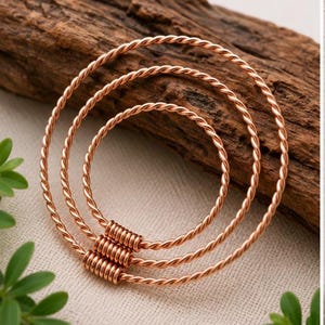 May include: Three copper-colored bangles with a twisted rope design. The bangles are arranged in a nested configuration, with a small section of tightly wound copper wire connecting them. The background features a piece of wood and green leaves.