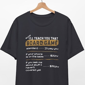May include: Black t-shirt with the text "I'LL TEACH YOU THAT BOARD GAME." The text includes the hourly rates for phone use and questions about uncovered topics. The shirt is hanging on a wooden hanger.