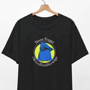 May include: Black t-shirt with a white circle graphic featuring a blue bird with a red eye. The text "Never Forget #WoodlandGrudge" is written around the bird.