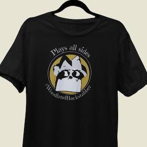 May include: Black t-shirt featuring a raccoon graphic inside a yellow circle, with the text "Plays all sides" and "#WoodlandBackstabber". The raccoon is white and black. The t-shirt is hanging on a black hanger.