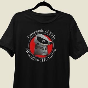 May include: Black t-shirt featuring a badger illustration inside a red circle, with the text "Crescendo of Pain" and "#WoodlandHistorian" in white. The badger is gray and white.