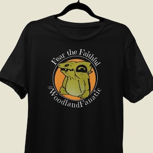 May include: Black t-shirt featuring a cartoon lizard inside an orange circle. The text reads "Fear the Faithful" above the lizard and "#WoodlandFanatic" below. The lizard is green and yellow.