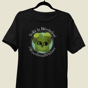 May include: Black t-shirt featuring a cartoon illustration of a green creature with the text "Voilà la Révolution!" and "#WoodlandWhiner" in white. The design is centered on the chest.