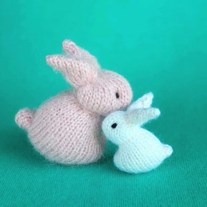 Op de afbeelding: Twee gebreide konijnen, een roze en een witte, knuffelen elkaar op een groene achtergrond. Het roze konijn is groter en heeft een meer gedefinieerd gezicht, terwijl het witte konijn kleiner is en een ronder gezicht heeft.