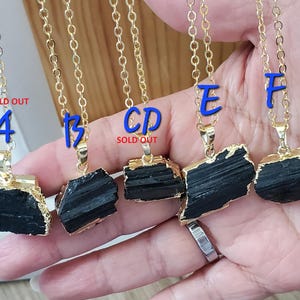 以下が含まれることがあります： ゴールドトーンのチェーンネックレス。生の黒トルマリンペンダント付き。ペンダントは金色の縁取りで、自然で粗い質感です。青い文字A、B、E、Fがチェーンに付いています。「SOLD OUT」の文字が2つのネックレスに表示されています。