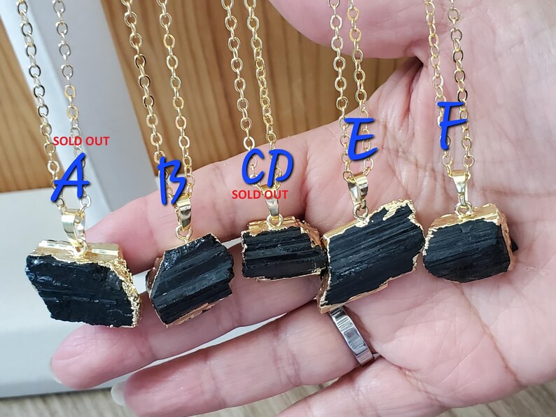以下が含まれることがあります： ゴールドトーンのチェーンネックレス。生の黒トルマリンペンダント付き。ペンダントは金色の縁取りで、自然で粗い質感です。青い文字A、B、E、Fがチェーンに付いています。「SOLD OUT」の文字が2つのネックレスに表示されています。