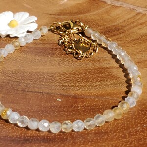 May include: A delicate gold chain bracelet with a white flower charm and clear and yellow gemstone beads.