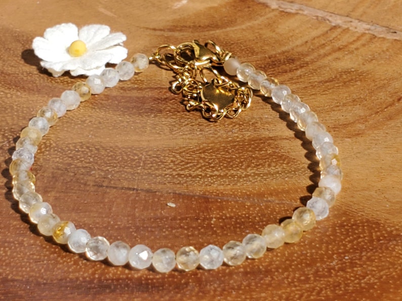 May include: A delicate gold chain bracelet with a white flower charm and clear and yellow gemstone beads.