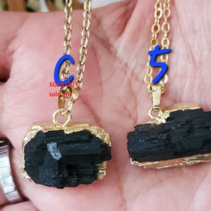 以下が含まれることがあります： 2つのゴールドカラーのチェーンネックレス。黒トルマリンのペンダントが付いています。ペンダントは、金色の縁取りのある、生の不規則な形をしています。「SOLD OUT」の文字が赤色で書かれたペンダントと、青色の数字「5」が書かれたペンダントがあります。
