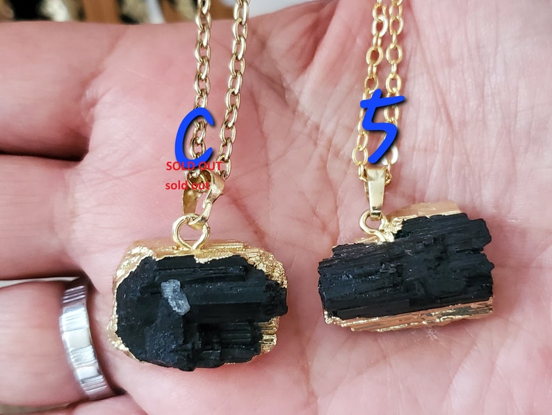 以下が含まれることがあります： 2つのゴールドカラーのチェーンネックレス。黒トルマリンのペンダントが付いています。ペンダントは、金色の縁取りのある、生の不規則な形をしています。「SOLD OUT」の文字が赤色で書かれたペンダントと、青色の数字「5」が書かれたペンダントがあります。