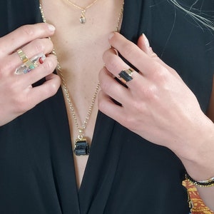 以下が含まれることがあります： ゴールドトーンのジュエリーコレクション。リングとネックレスがあり、宝石とクリスタルが特徴です。あるリングはカラフルな石が付いた透明なクリスタル、別のリングは黒い石です。黒いトップスが着用されています。