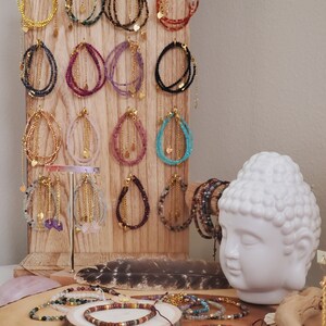 May include: A wooden display stand with rows of colorful beaded bracelets with gold accents. The bracelets are arranged in a grid pattern and feature various gemstone colors, including pink, purple, blue, green, and black. A white ceramic Buddha head sits in front of the display.
