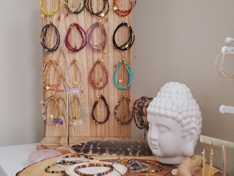 May include: A wooden display stand with rows of colorful beaded bracelets with gold accents. The bracelets are arranged in a grid pattern and feature various gemstone colors, including pink, purple, blue, green, and black. A white ceramic Buddha head sits in front of the display.