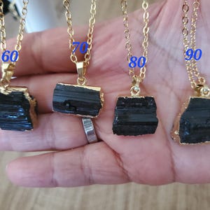 以下が含まれることがあります： 黒トルマリンのペンダントが付いた、4つのゴールドカラーのチェーンネックレス。各ペンダントは、金色の縁取りが付いた、ラフカットの長方形の石です。画像には、60、70、80、90の数字が青色で書かれています。