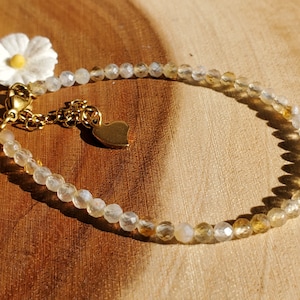 May include: A delicate bracelet made with clear and golden beads, featuring a small heart charm and a white daisy charm. The bracelet is on a wooden surface.