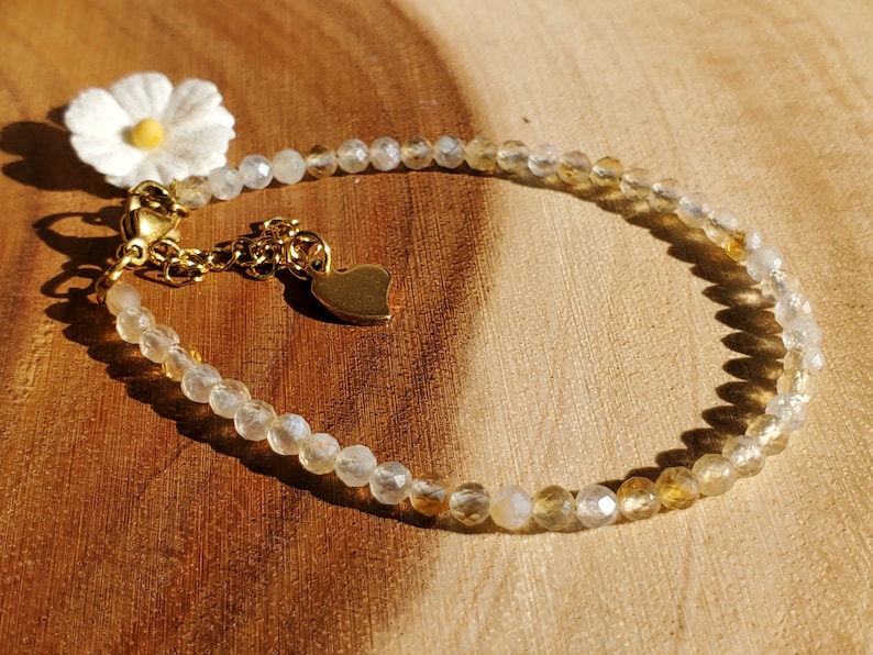 May include: A delicate bracelet made with clear and golden beads, featuring a small heart charm and a white daisy charm. The bracelet is on a wooden surface.