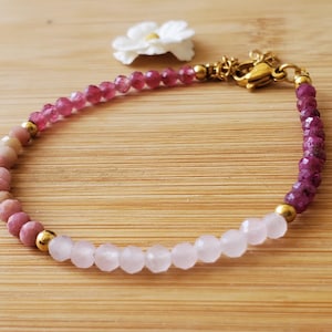 May include: A beaded bracelet featuring a mix of pink and purple gemstones. The bracelet has a gold-colored clasp and connectors. The gemstones include rose quartz, rhodochrosite, and tourmaline. A white flower is in the background.