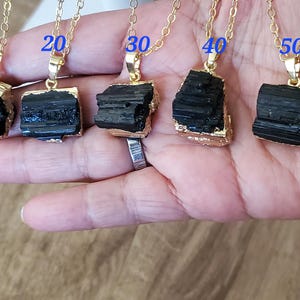 以下が含まれることがあります： 黒トルマリンのペンダントが付いた5つのゴールドトーンネックレス。各ペンダントは、金の縁取りが付いた、粗くカットされた長方形の石です。ネックレスは手の上に表示され、各ペンダントの上に10、20、30、40、50の数字が表示されています。