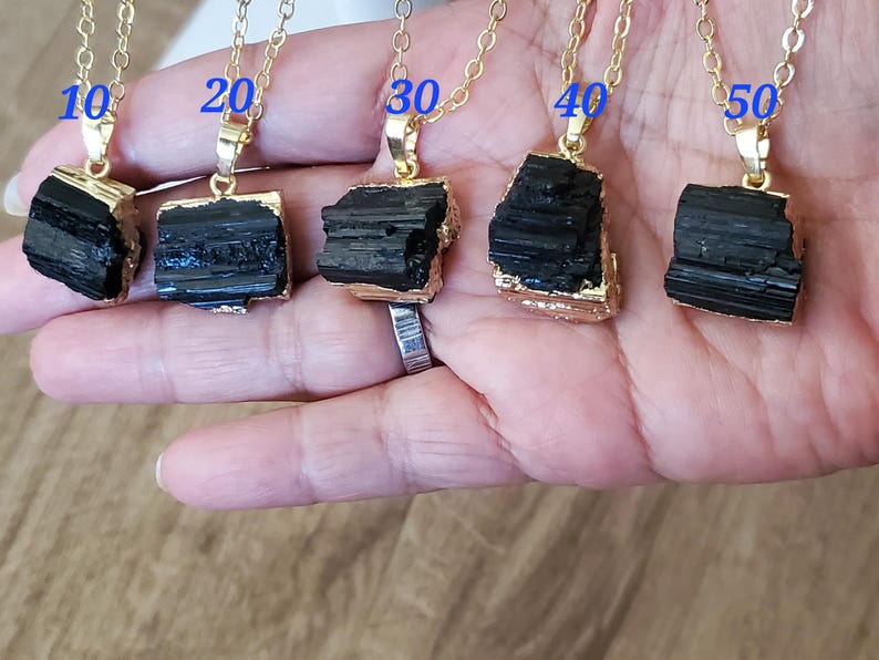 以下が含まれることがあります： 黒トルマリンのペンダントが付いた5つのゴールドトーンネックレス。各ペンダントは、金の縁取りが付いた、粗くカットされた長方形の石です。ネックレスは手の上に表示され、各ペンダントの上に10、20、30、40、50の数字が表示されています。