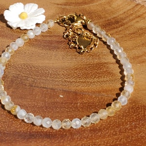 May include: A delicate gold chain bracelet with a white flower charm and clear and yellow beads.