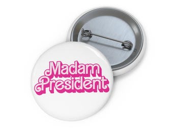 May include: A white button with a pink "Madam President" text in a retro font style.
