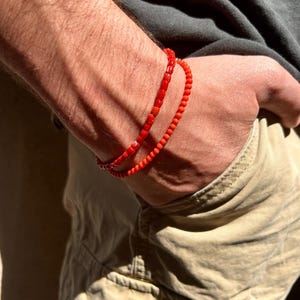 Könnte beinhalten: Zwei geschichtete Armbänder aus kleinen, leuchtend roten Perlen. Die Perlen sind in Größe und Form einheitlich und bilden ein durchgehendes kreisförmiges Design, das sich um das Handgelenk legt. Die helle rote Farbe hebt sich von der Haut ab und sorgt für eine schlichte und konsistente Ästhetik des Accessoires.
