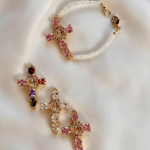 May include: A gold-toned bracelet featuring a cross pendant with pink teardrop-shaped stones and small white beads, alongside three separate cross pendants adorned with multi-coloured, clear, and pink teardrop-shaped crystals and central oval charms.
