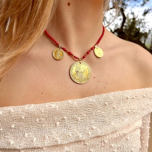 Puede incluir: Un collar de cuentas rojas con tres colgantes de monedas de oro. La moneda más grande presenta un perfil de una persona.