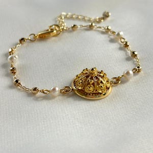 Może przedstawiać: Złota bransoletka z łańcuszka z małymi białymi perełkami i złotą zawieszką w kształcie kopuły. Zawieszka ma szczegółowy, skomplikowany wzór.