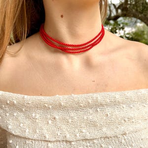 Może przedstawiać: Czerwony naszyjnik choker z wieloma rzędami koralik&oacute;w. Naszyjnik nosi osoba w białej bluzeczce z odkrytymi ramionami.