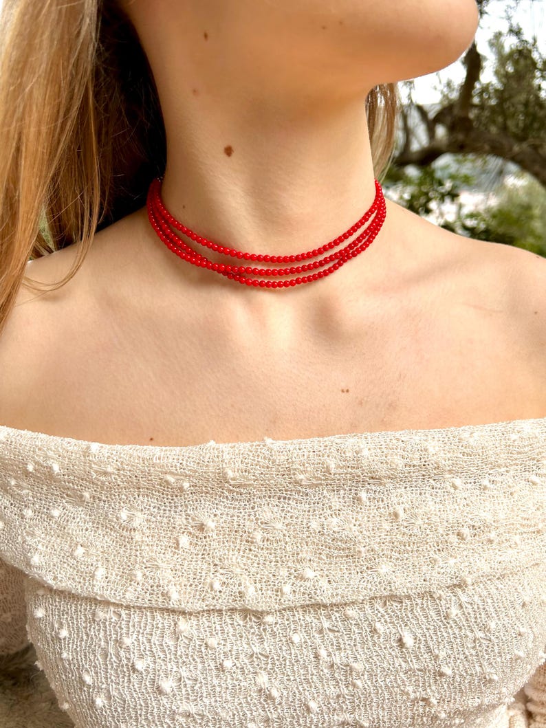 Może przedstawiać: Czerwony naszyjnik choker z wieloma rzędami koralik&oacute;w. Naszyjnik nosi osoba w białej bluzeczce z odkrytymi ramionami.