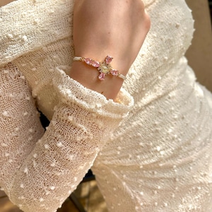 May include: A white beaded bracelet with a pink and gold cross charm. The cross has a clear center and pink stones around the edges.