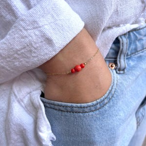 May include: A delicate gold chain bracelet featuring three small, round, vibrant orange-red beads centred on the chain. The bracelet is composed of a thin, polished gold-tone metal link chain that secures around the wrist with a simple clasp.