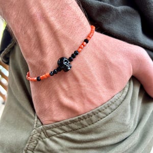 Op de afbeelding: Een kralenarmband met een centrale zwarte schedelbedel, geflankeerd door kleine, gefacetteerde zwarte kralen en een reeks feloranje cilindrische kralen. Het ontwerp wisselt af tussen de oranje en zwarte elementen voor een duidelijk contrast.