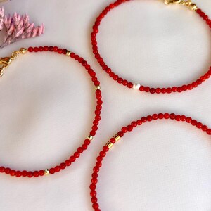 May include: Three delicate bracelets made of small, round, vibrant red beads. Each bracelet features a gold-toned metal lobster clasp and small gold-toned spacer beads. One bracelet includes a single white pearl accent, while others have gold-toned cylindrical accents.