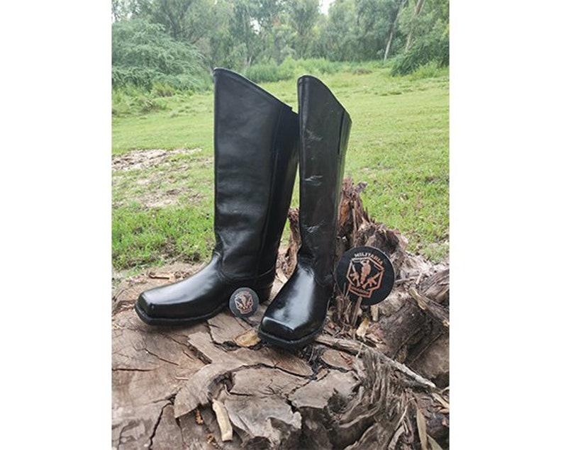 May include: A pair of black leather knee-high boots with a square toe. The boots are sitting on a tree stump.