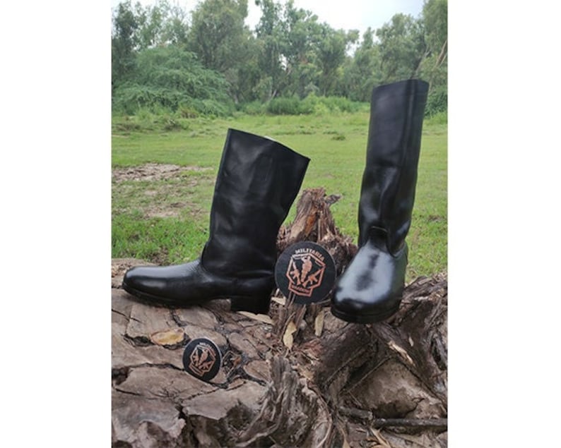 May include: A pair of black leather boots with a tall shaft. The boots are on a wooden surface with a brown background.